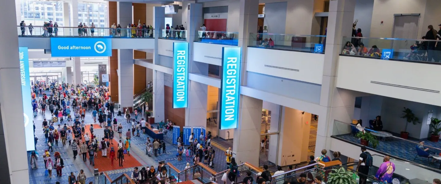 A large crowd of attendees gathers across multiple levels of a brightly lit convention center during an event, with prominent “Registration” signs and escalators connecting the floors.